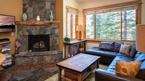A cozy living room with a stone fireplace, a large window with a forest view, a leather sofa, and a coffee table. - Mountain bikers dream near Gondola in Northstar (Truckee)