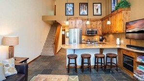 A modern kitchen with wooden cabinets, a central island with bar stools, a microwave, and a refrigerator. There is a staircase leading to another level. - Mountain bikers dream near Gondola in Northstar (Truckee)