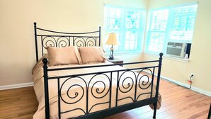 Een bed met een metalen frame en beige beddengoed, een nachtkastje met een lamp, en een raam met jaloezieën.