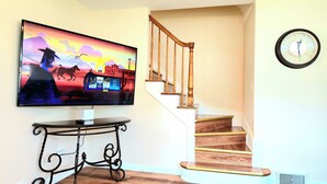 Een trap met houten treden en een houten leuning, een hangklok aan de muur en een flatscreen televisie waarop een film te zien is.
