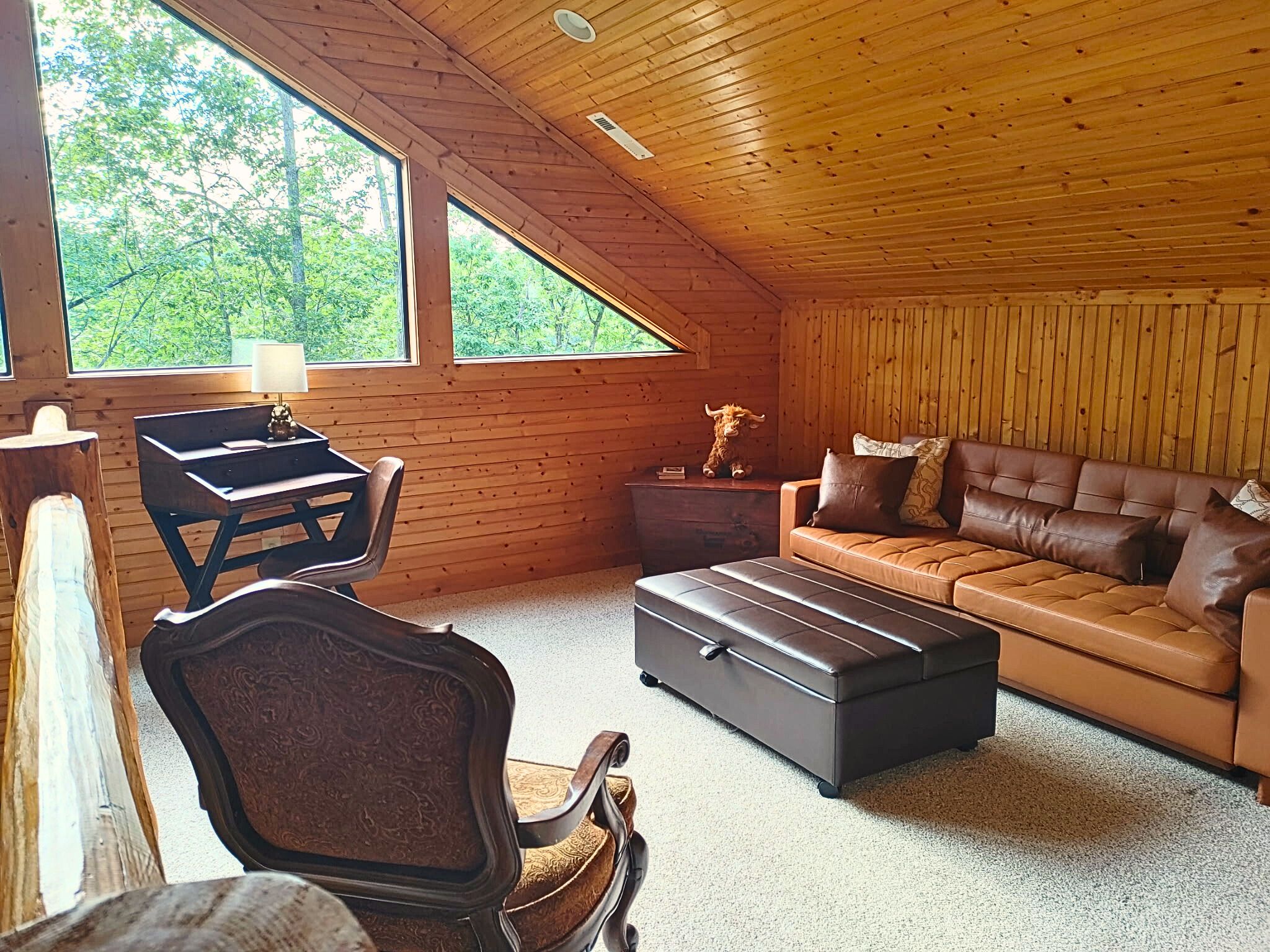 A cozy cabin interior with a wooden ceiling, a leather sofa, a wooden table, and a large window offering a view of trees.