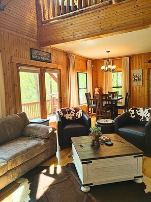 Interior acogedor de una cabaña con paredes de madera, un comedor y un letrero que dice 'Welcome to Neck of the Woods'.
