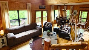 Una cabaña espaciosa con techo alto, paredes de madera, una zona de estar acogedora con sofá y mesa de centro, comedor y vistas al exterior a través de amplios ventanales.