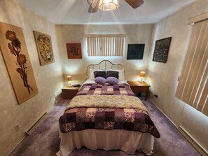 Ein Schlafzimmer mit einem großen Bett, zwei Nachttischen, einem Fenster mit Jalousien und verschiedenen Wandbildern.