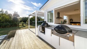 Een moderne buitenkeuken met een ingebouwde grill en een grote kookpot, gelegen op een houten terras met uitzicht op een aangelegde tuin en een huis op de achtergrond.