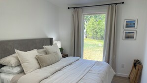 Un dormitorio con una cama grande, un cabecero, una mesita de noche con lámpara y vistas a un jardín a través de una ventana.