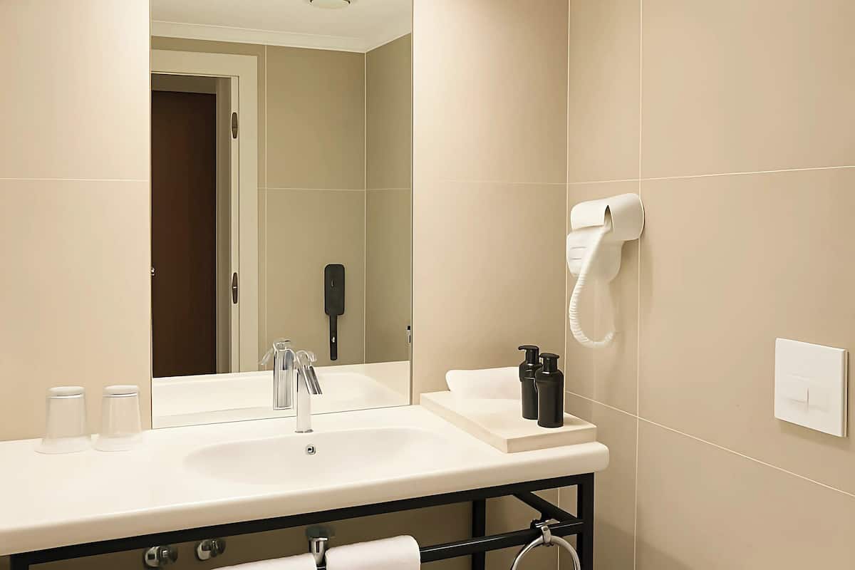 A modern bathroom with a white sink, mirror, and toilet. There is a hairdryer on the wall and a trash can next to the toilet.