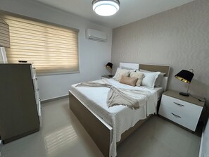Un dormitorio con cama, mesitas de noche, armario, ventana con persianas, unidad de aire acondicionado y una lámpara en el techo.