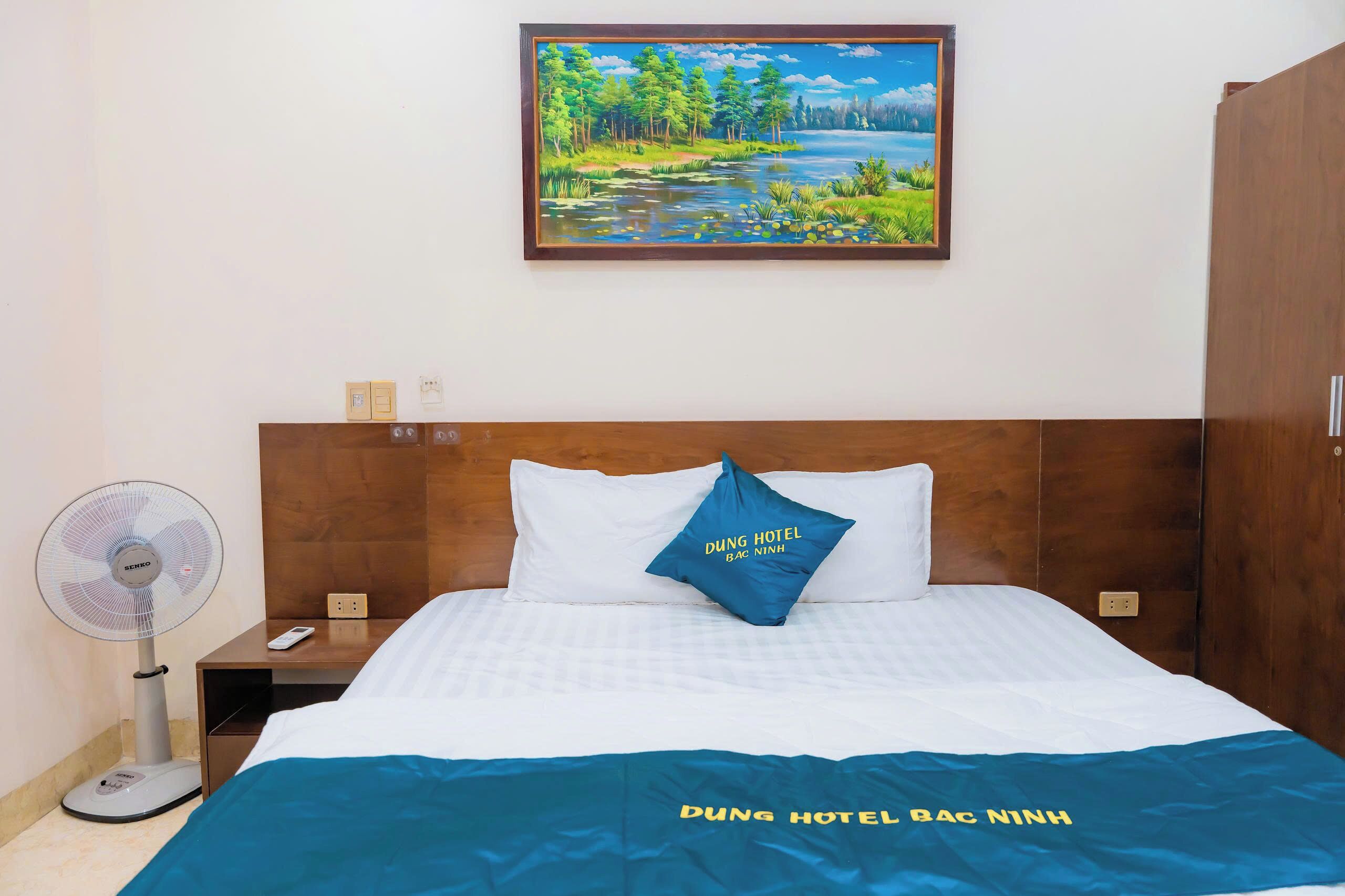 A hotel room with a bed, a nightstand, a wall-mounted fan, and a painting of a landscape.