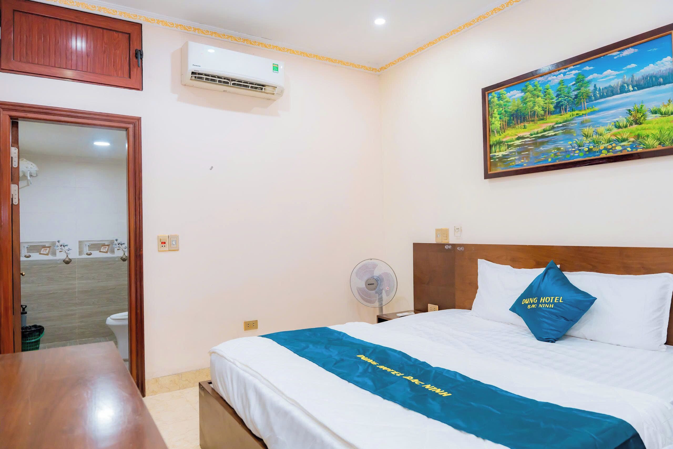 Una habitación de hotel con cama, ventilador, aire acondicionado, un cuadro y una puerta de madera.