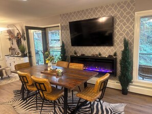 A dining area with a wooden table, leather chairs, a zebra-patterned rug, a fireplace with blue LED lights, and a mounted TV. -  Hot Tub! Charming 3-bedroom house in Estes Park- On the River! (Estes Park)