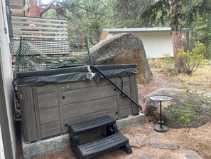 A hot tub with a wooden exterior, a small step stool, a metal table, and a white shed in a backyard with trees and a fence. -  Hot Tub! Charming 3-bedroom house in Estes Park- On the River! (Estes Park)