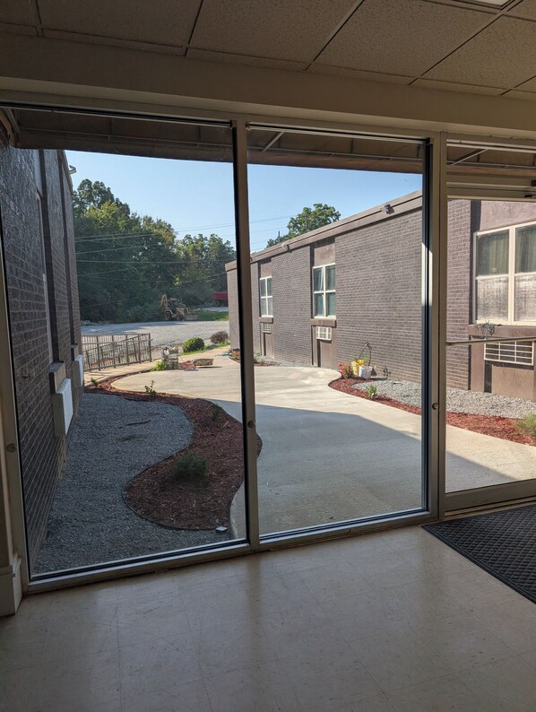 A view through a glass door of a building with a brick exterior, a paved courtyard, and a small garden area with mulch and plants.