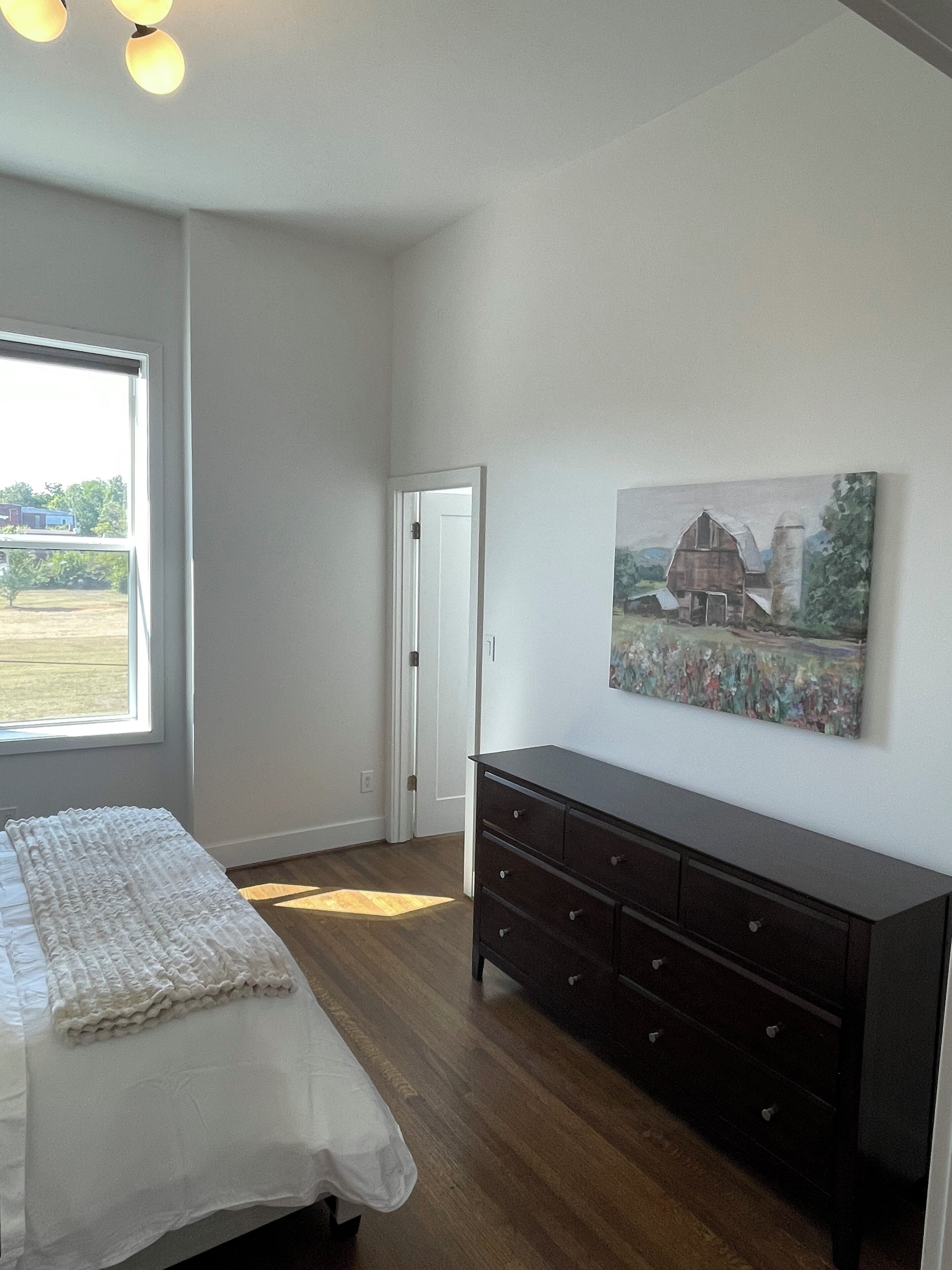 A bedroom with a bed, a dresser, a painting, and a window with a view of a field.