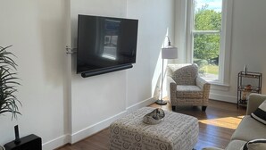 A living room with a flat-screen TV mounted on the wall, a patterned ottoman, a chair, and a large window providing a view of greenery outside.