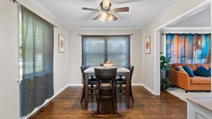 A dining area with a table and chairs, a ceiling fan, and large windows with curtains. - Escape by Beaches & Blue Angels (Pensacola)