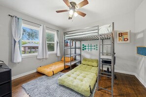 A bunk bed room with a ceiling fan, a window with curtains, a bunk bed with pillows, a desk, and a framed picture on the wall. - Escape by Beaches & Blue Angels (Pensacola)