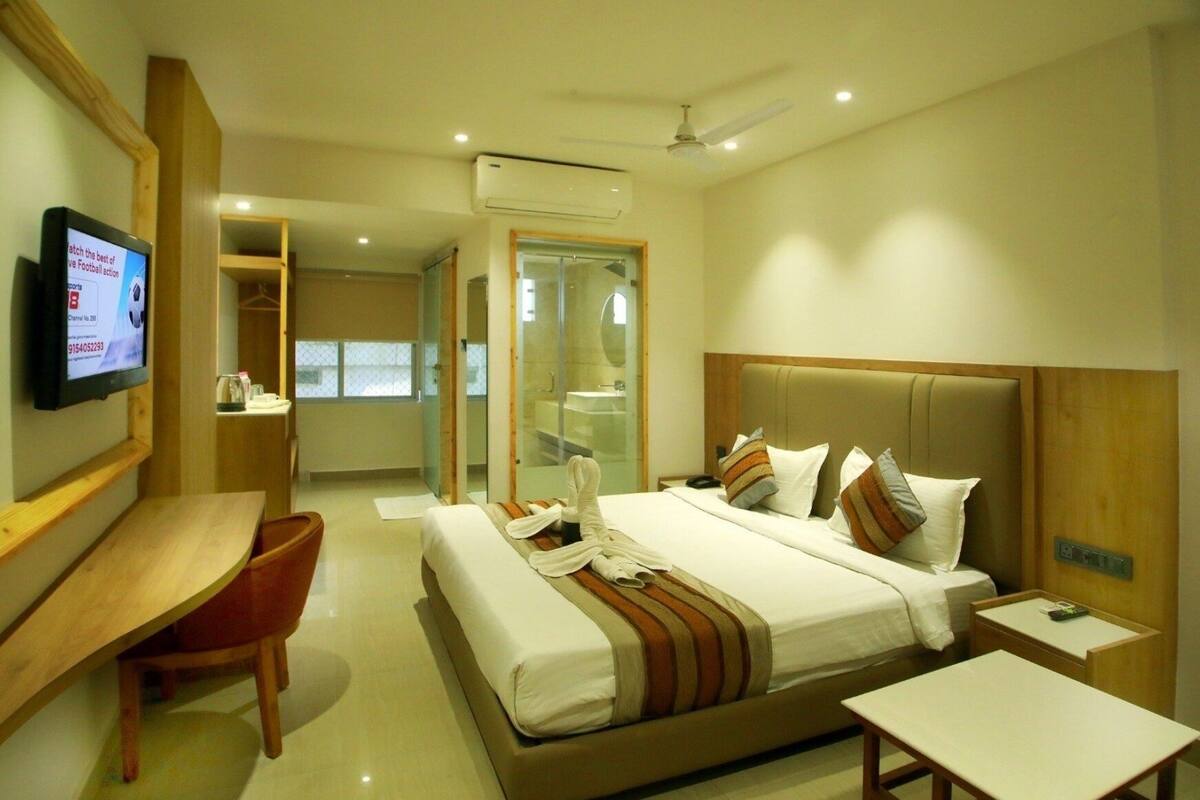 A hotel room with a large bed, a desk, a TV, and a bathroom visible through a glass door.