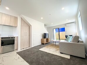 A modern living room with a grey sofa, a flat-screen TV on a wooden stand, and a large window offering a view of a blue sky and wooden fence.