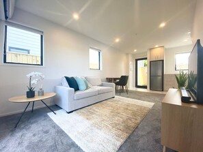 A modern living room with a sofa, a coffee table, and a TV. There is a kitchen area with cabinets and a refrigerator.