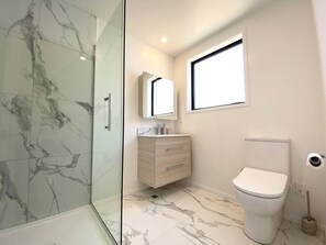 A modern bathroom with a glass shower enclosure, a marble-tiled floor, a white toilet, and a wooden vanity with a mirror. - Salisbury courtyard stay with fibre Wi-Fi in Christchurch (Christchurch)