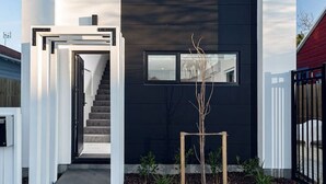 Une maison moderne à deux étages, dotée d’une façade sombre, d’un escalier apparent menant à l’entrée et d’un petit jardin agrémenté de plantes.