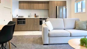 Une cuisine moderne avec des armoires en bois, un réfrigérateur en acier inoxydable et un four encastré. Le salon est aménagé avec un canapé gris et un fauteuil noir.