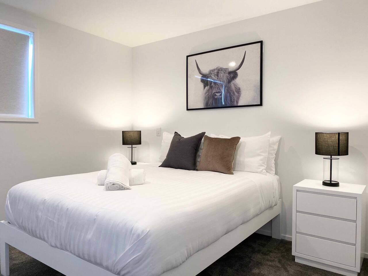 A hotel room with a large bed, white bedding, two bedside tables with lamps, a framed artwork on the wall, and a window with blinds.