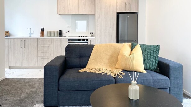 Una sala de estar moderna con un sofá azul oscuro, una mesita de centro redonda y una cocina con armarios de madera clara y un refrigerador negro.