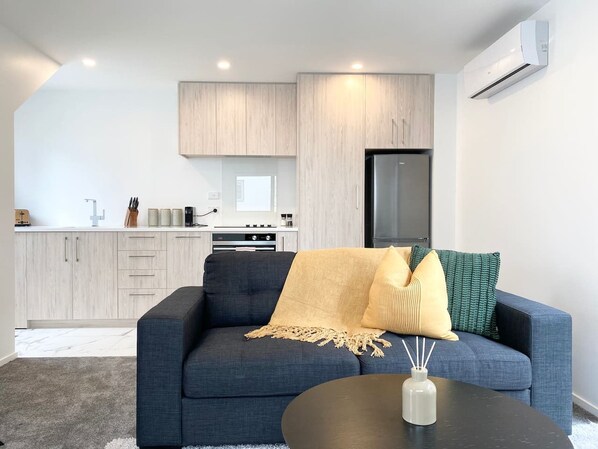 A modern living room with a dark blue sofa, a small round coffee table, and a kitchen area with light wood cabinets and a black refrigerator.