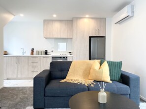 A modern living room with a dark blue sofa, a small round coffee table, and a kitchen area with light wood cabinets and a black refrigerator.