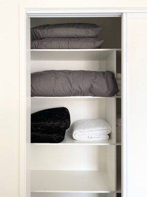 Ein Schrank mit drei Ablagen, auf denen ein gefaltetes weißes Handtuch, ein schwarzes Samtkissen und zwei graue Kissen liegen.