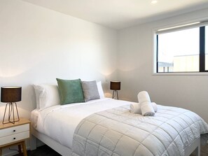 Ein ordentlich bezogenes Bett mit weißer Bettdecke und Kissen, eine Nachttischlampe und ein Fenster mit Jalousien.