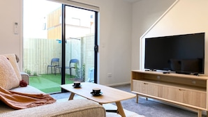 Ein Wohnzimmer mit einer Couch, einem Couchtisch, einem Fernseher auf einem Holzständer und einer Glastür zum Patio mit Stühlen und einem kleinen Garten.