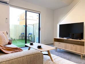 Ett vardagsrum med en soffa, ett soffbord, en tv på ett träställ och en skjutdörr i glas som leder ut till en uteplats med stolar och en liten trädgård.