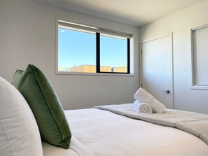 Una cama bien hecha con sábanas blancas y un cojín verde, una ventana con persianas, y una puerta en una habitación con paredes blancas.
