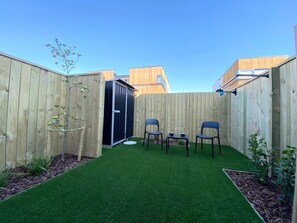 Un jardín trasero con césped artificial verde, dos sillas azules, una mesita y una valla de madera.