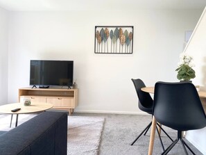 A modern living room with a flat-screen TV, a wooden entertainment unit, a round coffee table, a black sofa, a dining table with black chairs, and a decorative wall piece. - Salisbury courtyard retreat with a workspace in Christchurch (Christchurch)