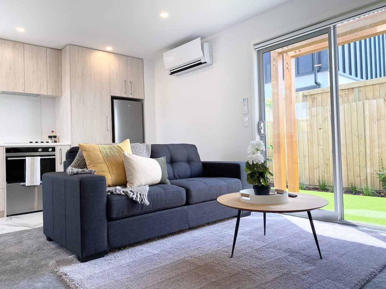 A modern living room with a dark sofa, a round coffee table, and a kitchen area with wooden cabinets and an air conditioning unit.