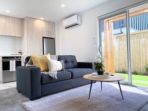 A modern living room with a dark sofa, a round coffee table, and a kitchen area with wooden cabinets and an air conditioning unit. - Salisbury courtyard retreat with a workspace in Christchurch (Christchurch)