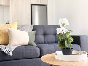 Ein modernes Wohnzimmer mit einer grauen Couch, einem runden Holztisch und einem Blumentopf mit weißen Blüten.