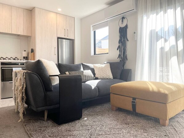 A modern living room with a grey sofa, a black ottoman, and a beige ottoman. There is a kitchen area with wooden cabinets and a stainless steel refrigerator. A dreamcatcher hangs on the wall, and a window with sheer curtains allows natural light into the room.