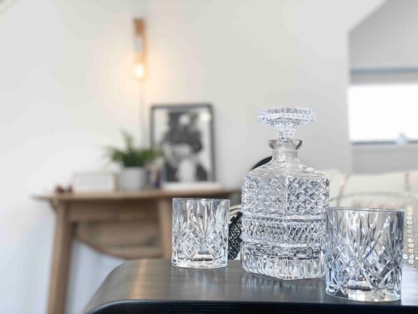 Un decantador de vidrio transparente y dos vasos sobre una mesa en una habitación bien iluminada, con una chimenea y un cuadro enmarcado al fondo.