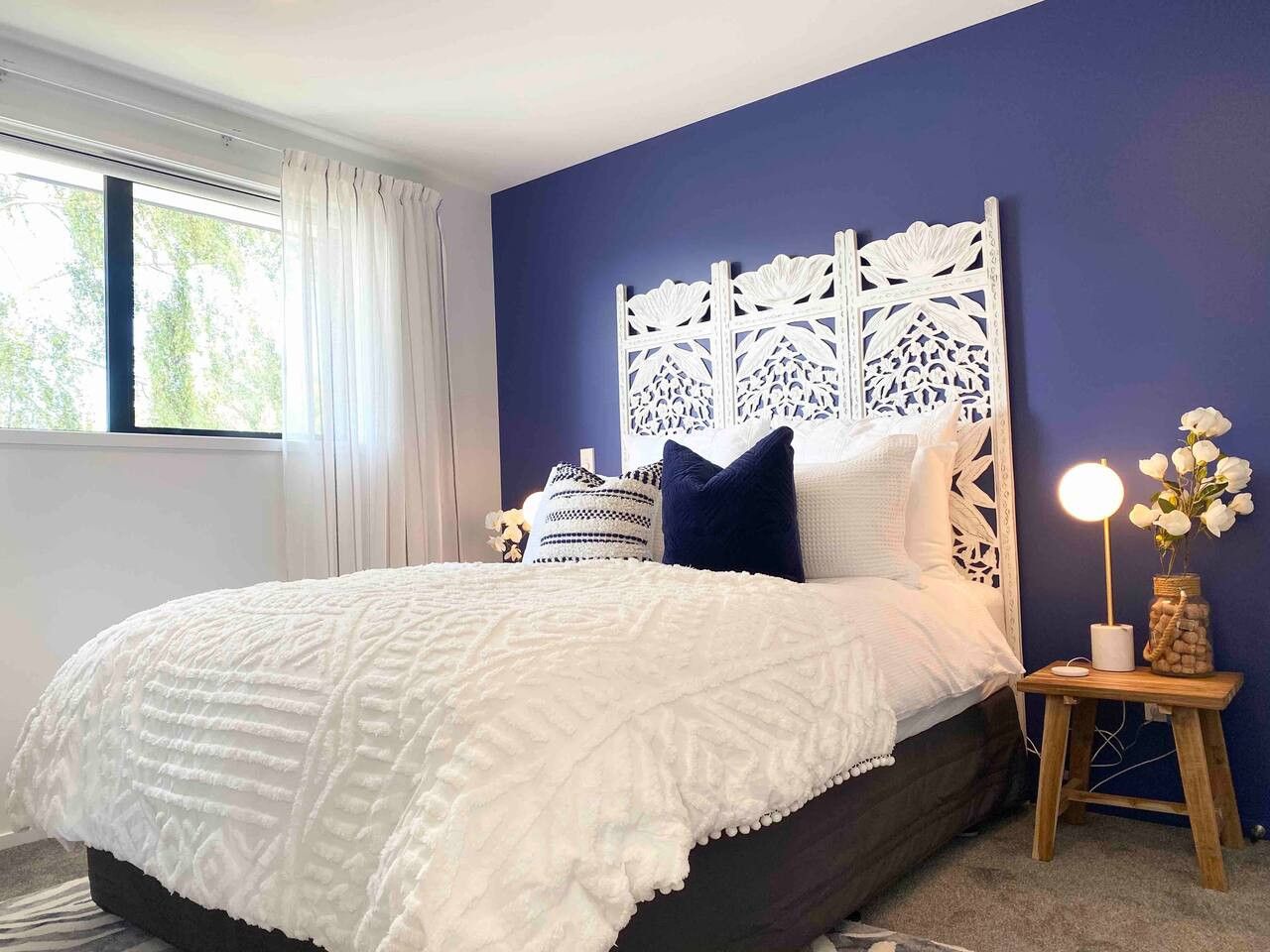 Une chambre à coucher avec un couvre-lit blanc, une tête de lit décorative, une table de chevet avec une lampe et des fleurs, et une fenêtre avec des rideaux.