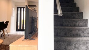 A modern living room with a flat-screen TV on a wooden stand, a dining area with chairs, and a staircase with dark carpeted steps.