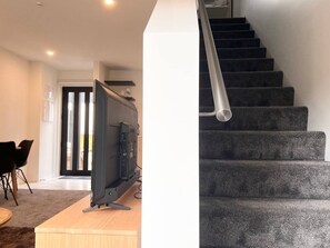 Ein modernes Wohnzimmer mit Flachbildfernseher auf einem Holzboden, einem Essbereich mit Stühlen und einer Treppe mit dunkel bezogenen Stufen.
