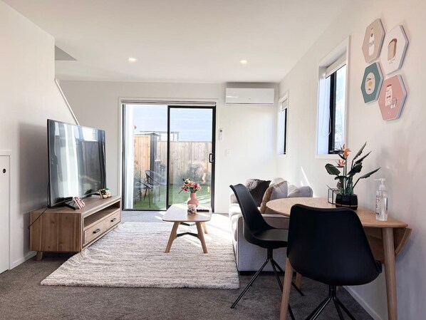 A modern living room with a TV, a dining area with a table and chairs, and a large sliding glass door leading to a backyard.