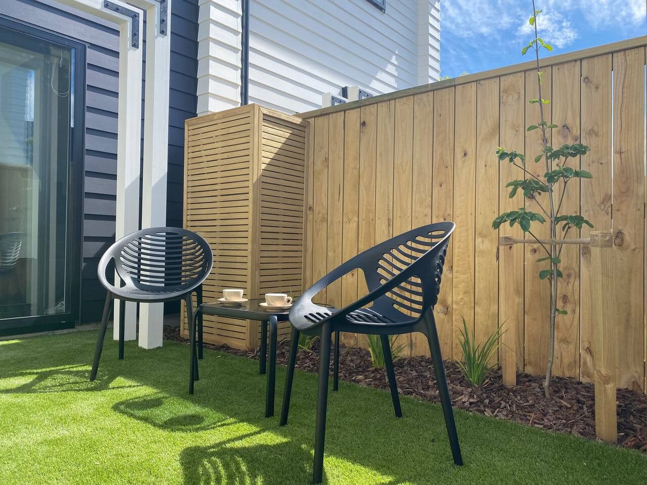An outdoor patio with two chairs and a table, a wooden fence, and a small plant.