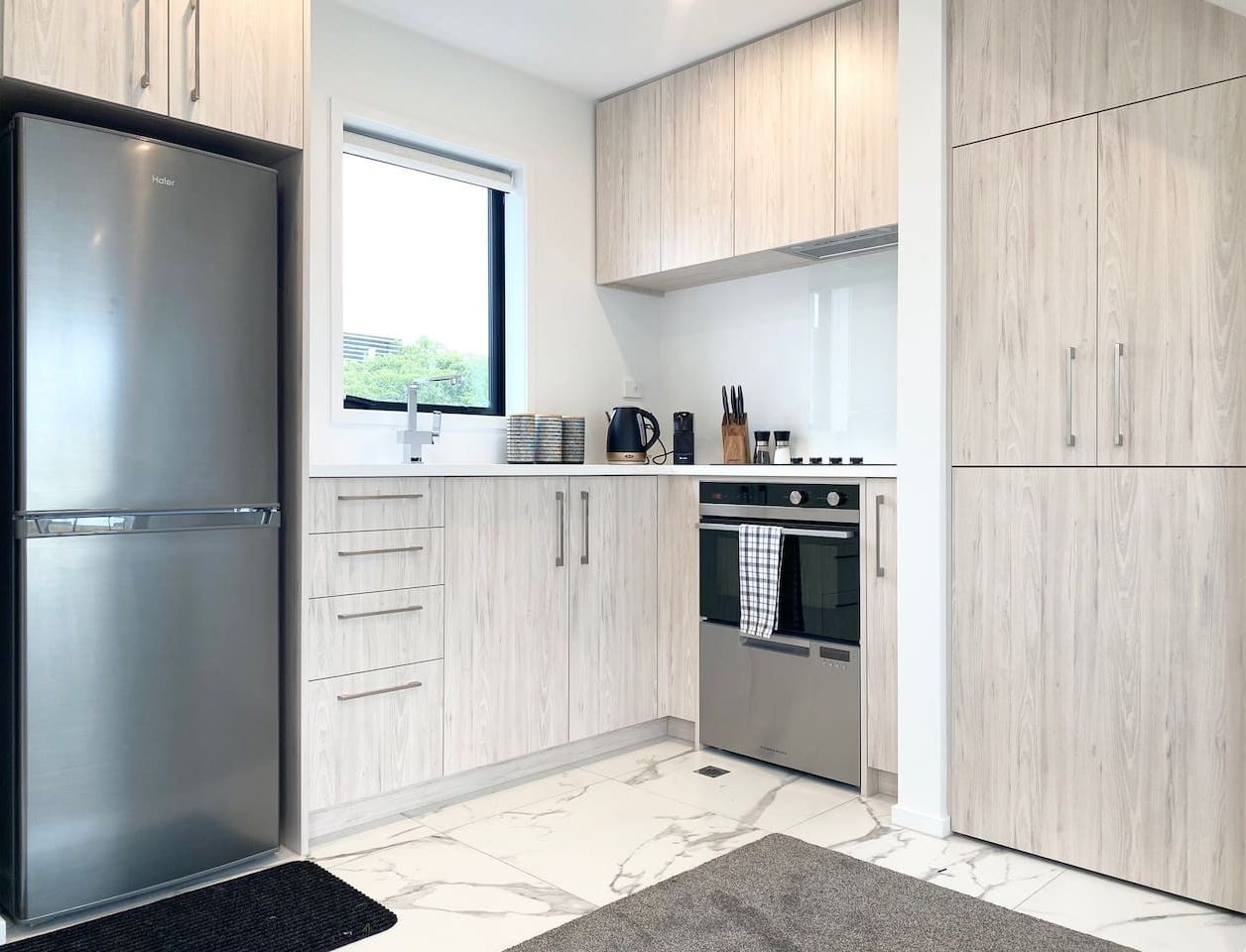 A modern kitchen with light wood cabinets, stainless steel appliances, and a large window providing natural light.