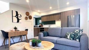 Uma sala de estar moderna com um sofá cinzento, uma mesa de centro redonda com plantas, uma mesa de jantar com cadeiras e uma cozinha ao fundo.